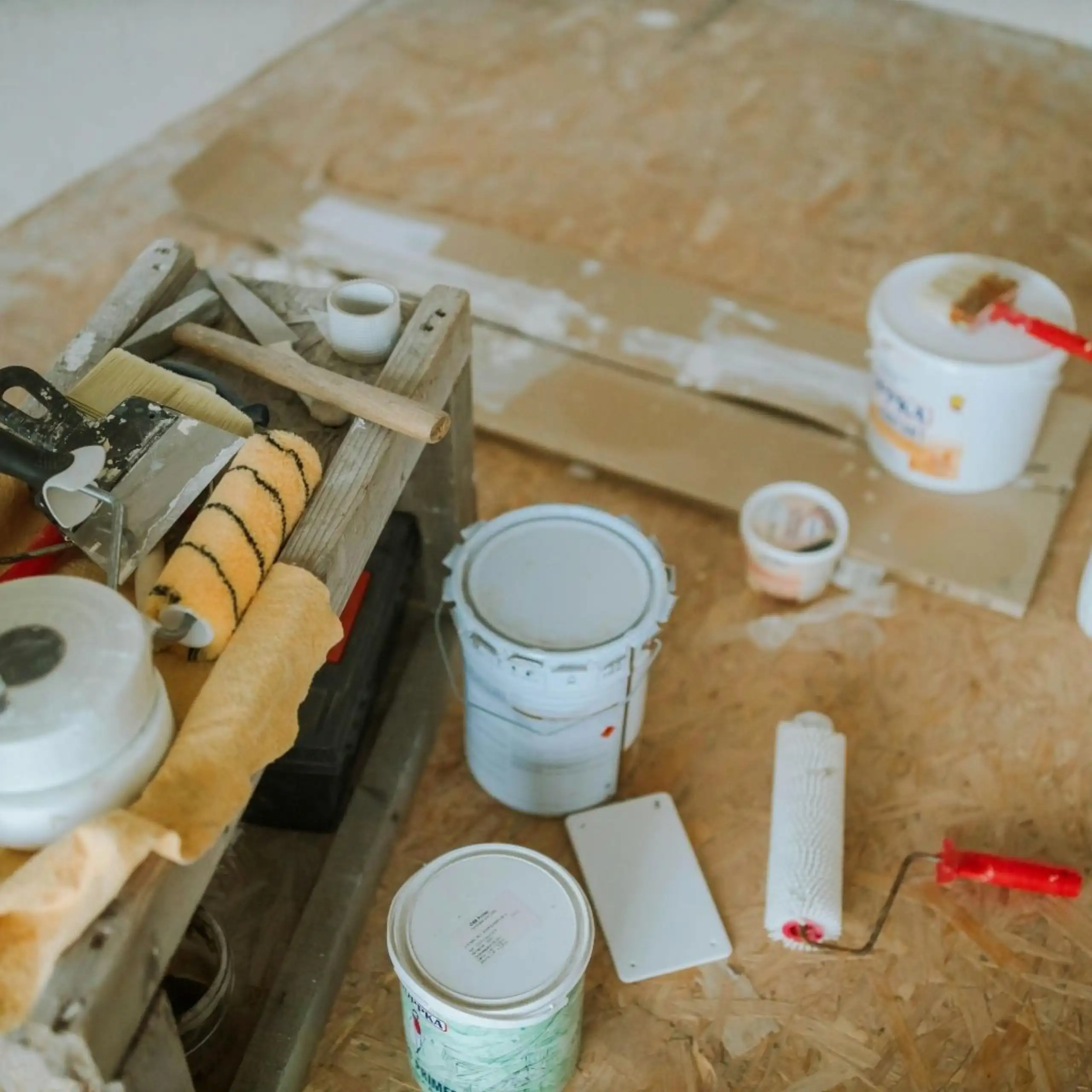 Home remodeling in progress with paint buckets, roller, and putty knives on a plywood floor.